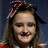 A person with long brown hair wearing a red and white headband and red clothing, smiling against a dark blue background.