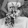 Black-and-white static image of a person with a large bouquet of flowers held close to the face, wearing a light-colored shirt and a wide-brimmed hat, with a blurred background of foliage.