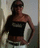 Pixelated image of a person with dark hair tied back, wearing a black sleeveless top and light-colored pants, standing against a muted background.