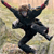 Pixelated figure in black attire with red accents on arms, arms raised high, legs bent in dynamic pose against blurred background.
