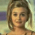 Static pixelated image of a person with shoulder-length brown hair, light skin, and a neutral expression, wearing a dark strap top against a blurred background.