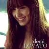 A close-up of a person with long brown hair and bangs smiling, wearing a light-colored top with a pink shoulder detail, against a blurred background with text overlay.