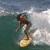 A person in a dark top and light pants rides a yellow surfboard on a greenish wave with white foam under a gray sky.