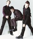 Static image of three men in dark formal attire posing against a light background, with one seated and two standing on either side.