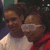 Two individuals in close proximity, one wearing a white shirt and the other in red, with dark hair and a blurred background.