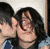 Pixelated static image of two people with dark hair in close proximity, one person partially visible on the left side and another more prominent figure on the right with long black hair, both wearing dark clothing against a light background.