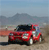 Red vehicle with dark roof and white accents on wheels, positioned on a sandy surface under a light blue sky with distant mountains.