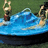 Two figures in a blue watercraft on green background; one figure raises arms, the other holds an object.