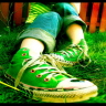 Static close-up of a person's foot wearing green high-top sneakers with white laces and yellow soles, positioned on grassy ground with blurred wooden fence background.