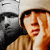 Pixelated static image of a person with light brown hair wearing a beige jacket and white shirt, positioned next to a grayscale figure in dark clothing against a neutral background.