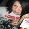 A person with long dark hair wears a white shirt with red text and a black-and-white graphic, holding a white object with red text.