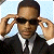Pixelated image of a person in a black suit and white shirt adjusting dark sunglasses with both hands against a light blue background.