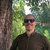 Pixelated image of a person wearing dark sunglasses and a dark shirt, standing outdoors with green foliage and a tree trunk in the background.