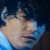 Pixelated still image of a figure with light skin tone, dark hair, and blue accents against a dark background.
