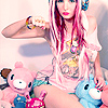 A person with long pink hair wearing a pink off-shoulder top and colorful accessories poses with plush toys.