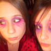 Two people with brown hair and pink-purple eyeshadow appear in a close-up, wearing red patterned clothing against a dark background.