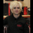 Pixelated image of a person with short blonde hair in dark clothing, surrounded by red and brown background elements, with bold red text overlays on black and gray