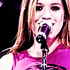 A person with light brown hair wearing a sleeveless pink top sings into a black microphone with a silver band, set against a dark background.