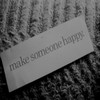 A grayscale image shows a rectangular card with the text 'make someone happy' on a textured fabric background.