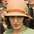 A pixelated close-up of a person wearing a light brown wide-brimmed hat with an orange band, dark curly hair, red lips, and a light green garment, with blurred figures in the background.