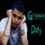 Pixelated static image of a person with dark hair wearing a white sleeveless shirt, holding hands near their face against a black background with green text reading "Green Day" on the right side.