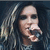 Pixelated close-up of a person with long light-colored hair holding a microphone against dark background.