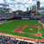 Pixelated static image of a baseball field with green grass, brown dirt, and distant city buildings under a light blue sky.