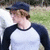 Pixelated static image of a person wearing a dark blue cap and dark blue-and-white raglan shirt, standing outdoors with blurred green and brown background.