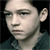 Pixelated close-up of a person's face with dark hair and a dark collared shirt against a black background, rendered in low-resolution grayscale tones.