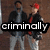 Two men stand side-by-side against a brown brick wall; one wears a red cap and black jacket over gray shirt, the other dark jacket over light blue shirt.