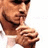Pixelated close-up of a person with dark hair and beard, wearing sunglasses, holding index finger to lips in silence gesture against white background.