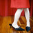 Pixelated image of a figure in white tights and red top standing on a brown floor with dark baseboard.