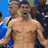 A shirtless man with short dark hair stands in front of a blurred crowd, mouth open as if shouting or cheering.