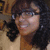 A person with dark curly hair and glasses smiles in a static image, wearing a light-colored top against a blurred background.