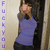 A person in a purple top and dark pants stands against a light background, making a hand gesture with the right hand raised near the face, while vertical purple text appears on the left side of the frame.
