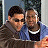Two pixelated figures in close proximity, one wearing dark sunglasses and a brown jacket, the other in a blue shirt with a white collar, against a blurred background.