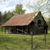 Pixelated static image of a small house with a brown roof, dark walls, and a grassy field under a light blue sky with clouds.