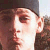 Pixelated close-up of a person's face with brown hair, wearing a dark cap and dark clothing, against a light background.