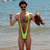 A pixelated figure in a green one-piece swimsuit stands on a beach with arms raised, holding objects in each hand against a blurred water and sand background.
