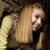Pixelated image of a person with long light brown hair wearing a yellow top, holding a dark object near their face against a dark background.