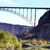 Static pixelated image of a suspension bridge spanning a body of water with rocky cliffs and greenery below under a light blue sky.