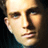 Pixelated close-up of a person's face with light skin tone, dark hair, and dark clothing against a dark background.