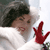 A person with dark curly hair and red lipstick wears a white garment, holding a red hand-shaped object near their face.
