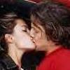 Two people in close physical proximity with lips touching, one wearing a red garment, the other in dark clothing, against a blurred background.