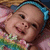 A pixelated close-up of a smiling child with light skin, dark eyes, and brown hair tied back by a blue headband, wearing a colorful patterned top.