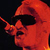 Pixelated red and orange cartoon character with dark sunglasses, a raised finger gesture, and a hooded silhouette against black background.