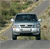 Static pixelated image of a light-colored vehicle with dark windows and headlights, positioned on a road with blurred green and brown background elements.