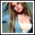 A pixelated static image of a person with long brown hair wearing a light blue top, set against a blurred background.