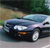 Static image of a dark blue sedan with a light blue roof driving on a road, surrounded by greenery and a blurred background.