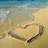 A heart shape drawn in wet sand on a beach with gentle waves lapping at the shore, viewed from above.